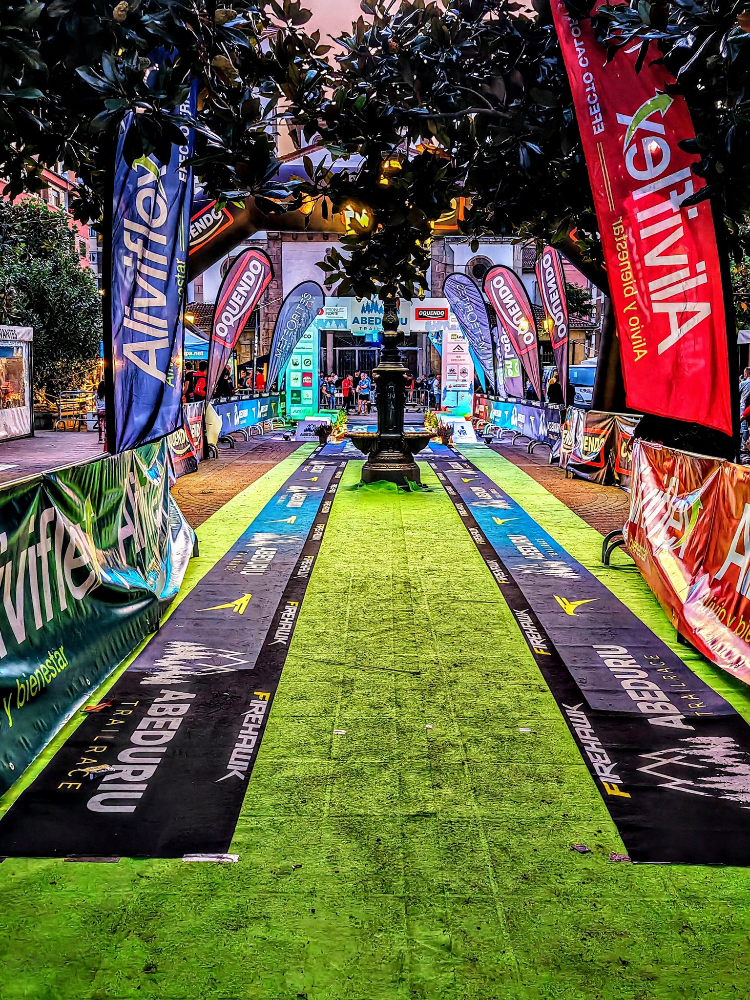 Recta de meta al anochecer, con alfombra verde, banderolas de los patrocinadores y una fuente en el centro de la plaza