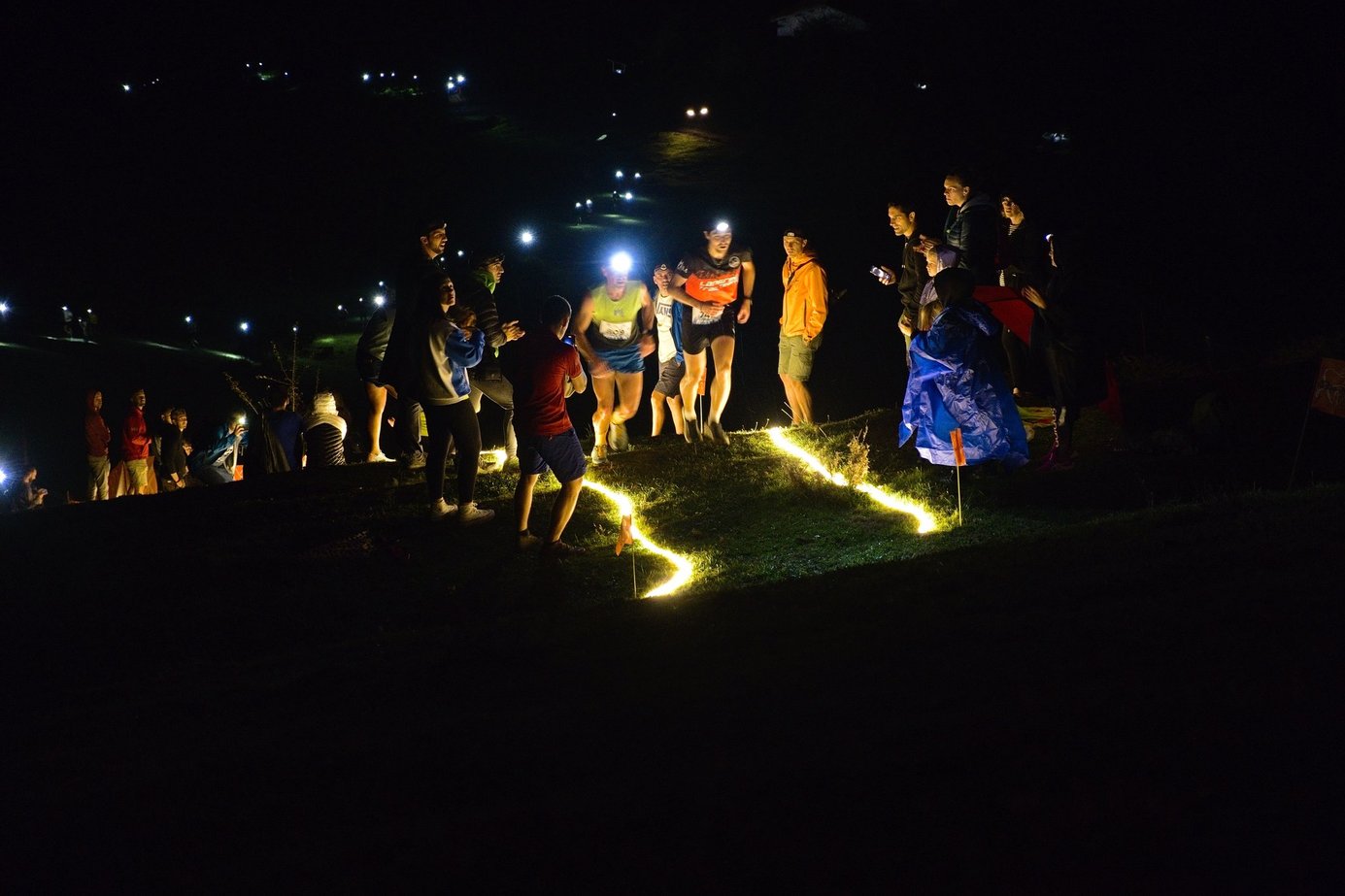 Corredores con linterna frontal pasan de noche por un tramo marcado con luces, entre el público